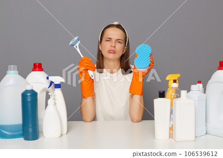 Sad upset unhappy woman posing at workplace with cleaning detergents holding sponge and brush crying looking at camera with pout lips isolated over gray background. 106539612