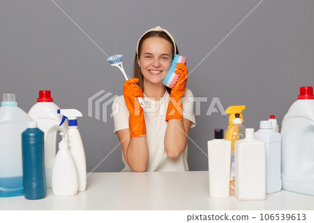 Cheerful positive woman posing at workplace with cleaning detergents isolated over gray background sitting with brush in hand holding sponge near ear pretends talking phopne. 106539613