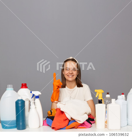 Delighted satisfied woman posing at workplace with cleaning detergents isolated over gray backgroun doing laundry pointing up at copy space for promotion. 106539632
