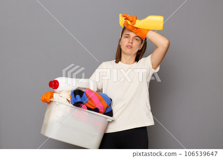 Housewife takes care of clothing. Domestic appliances aid in housework. Tired exhaused woman wearing whote t-shirt with laundry bowl and detergent isolated over gray background Housewife takes care of clothing. Domestic appliances aid in housework. Tired exhaused woman wearing whote t-shirt with laundry bowl and detergent isolated over gray background 106539647