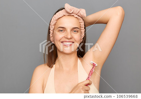 Hygiene skin body care. Hair removing. Satisfied young woman with hair band using razor, shaving armpit posing isolated over gray background. Hygiene skin body care. Hair removing. Satisfied young woman with hair band using razor, shaving armpit posing isolated over gray background. 106539668