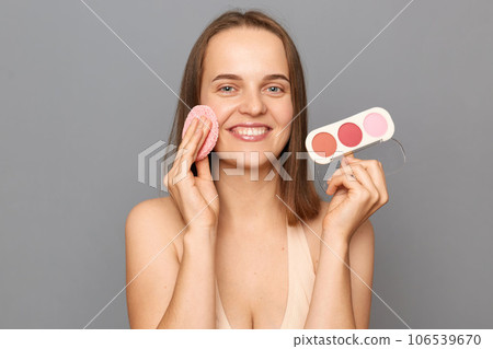 Skin care and beauty. Daily makeup. Smiling attractive young woman with brown hair holding sponge and powder posing isolated over gray background. Skin care and beauty. Daily makeup. Smiling attractive young woman with brown hair holding sponge and powder posing isolated over gray background. 106539670