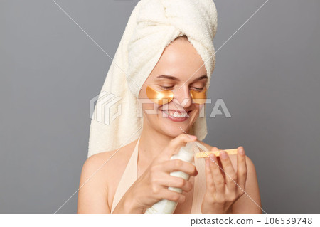 Cosmetic procedures. Skin care. Happy joyful young woman wrapped head in towel with patches under eyes using lotion to take care of face skin posing isolated over gray background. 106539748