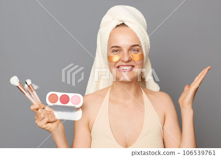 Beauty procedures. Makeup. Smiling woman with patches under eyes wrapped in towel advertising powder and brush raised her arm isolated over gray background. 106539761