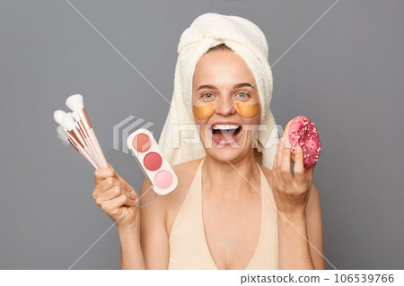 Excited attractive young woman with patches under eyes wrapped in towel posing isolated over gray background doing daily makeup and having tasty sweet snack. Excited attractive young woman with patches under eyes wrapped in towel posing isolated over gray background doing daily makeup and having tasty sweet snack. 106539766