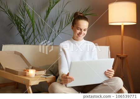 Smiling joyful attractive woman with bun hairstyle in white sweater looking at laptop screen sitting on sofa at home having video call watching movie. 106539771