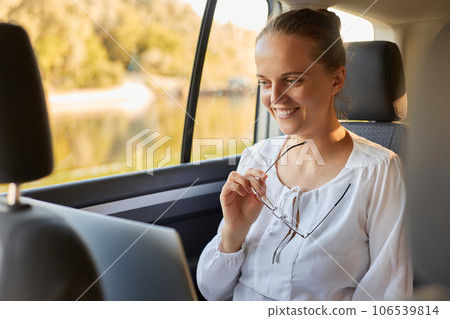 Beautiful confident woman working on laptop while sitting in the car having video call looking at display with smile holding eyeglasses while riding to work. Beautiful confident woman working on laptop while sitting in the car having video call looking at display with smile holding eyeglasses while riding to work. 106539814