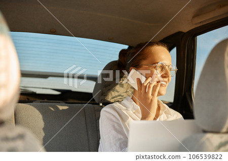 Joyful carefree young woman wearing white shirt and glasses talking smartphone sitting in car looking in auto widow with happy expressing pleasant conversation. Joyful carefree young woman wearing white shirt and glasses talking smartphone sitting in car looking in auto widow with happy expressing pleasant conversation. 106539822