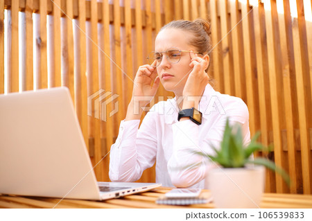 Tired concentrated woman wearing white shirt sitting in outdoor cafe working on laptop massaging templas has headache thinking about new job tasks. 106539833