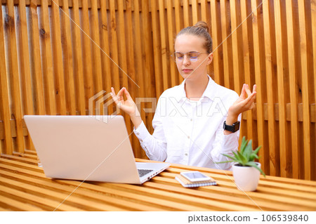 Relaxed overworked woman wearing white shirt sitting in outdoor cafe working on laptop trying to calm down, meditating practicing yoga, breathing. 106539840