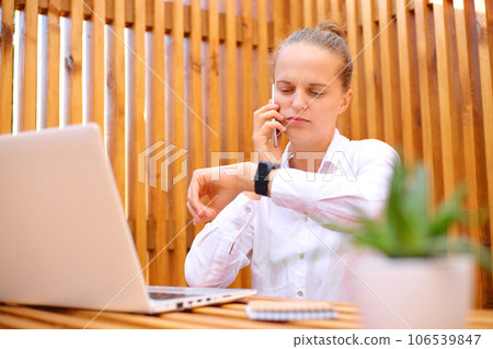 Busy woman wearing white shirt sitting in outdoor cafe working on laptop talking on mobile phone looking at her wristwatch worried about deadline. Busy woman wearing white shirt sitting in outdoor cafe working on laptop talking on mobile phone looking at her wristwatch worried about deadline. 106539847