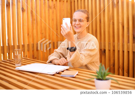 Smiling attractive woman wearing beige sweater sitting in outdoor cafe working on laptop having video call online while having rest from freelance job having enjoyable expression. Smiling attractive woman wearing beige sweater sitting in outdoor cafe working on laptop having video call online while having rest from freelance job having enjoyable expression. 106539856
