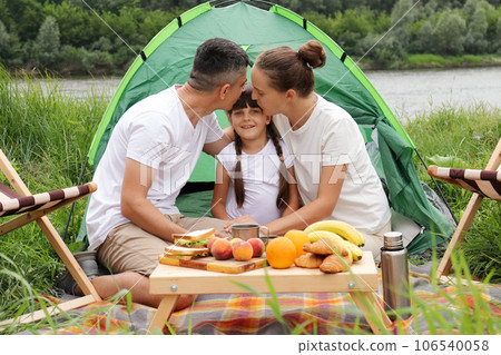 Camping near the river in nature park. Family at vacation. Couple with daughter resting. Parents kiss=sing daughter. People enjoying vacation. 106540058
