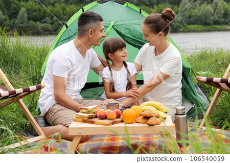 Family resting at riverside. Happy camping near the river in nature park, Couple with daughter sitting near tent. Weekends in the open air. 106540059