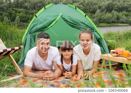 Nature vacation. Family trip. Camping trip, Mother daughter father lying on grass and blanket looking at camera, enjoying resting near the lake or river. Nature vacation. Family trip. Camping trip, Mother daughter father lying on grass and blanket looking at camera, enjoying resting near the lake or river. 106540066