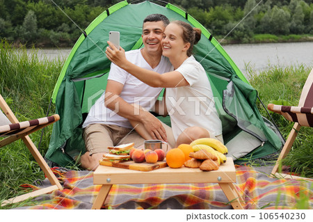 Happy woman and man wearing casual clothing sitting at tent near the river making selfie on smart phone enjoying resting in nature, 106540230