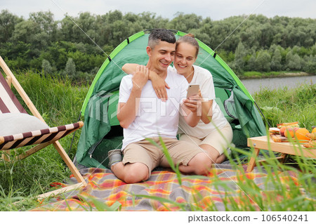 Beautiful woman and handsome man wearing casual clothing sitting at tent near the river using mobile phone, checking networks while having rest in nature. 106540241