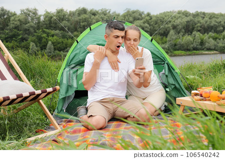 Shocked astonished woman and man wearing casual clothing sitting at tent near the river, couple sing cell phone, looking at display with surprised face. 106540242