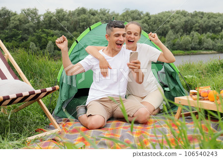 Excited joyful woman and man wearing casual clothing sitting at tent near the river, using mobile phone, clenched fists, rejoicing, having fun, reading n good news, betting and winning. 106540243