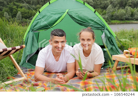 Resting outdoor. Smiling woman and man wearing casual clothing laying at tent near the river looking at camera, resting, camping at riverside. 106540244