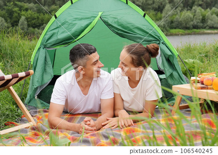 Joyful couple in nature, woman and man wearing casual clothing laying at tent near the river, happy vacation, romantic weekend, enjoying communication with each other. 106540245