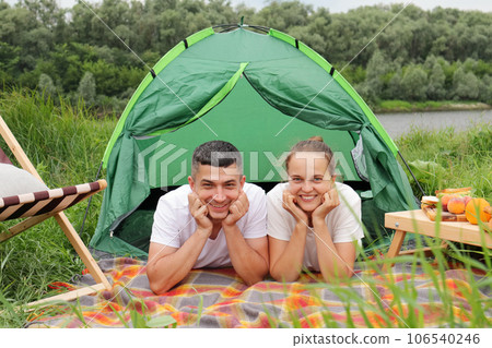 Young caucasian couple woman and man wearing casual clothing laying near tent by the river, having vacation together, romantic weekend in nature,looking at camera. Young caucasian couple woman and man wearing casual clothing laying near tent by the river, having vacation together, romantic weekend in nature,looking at camera. 106540246