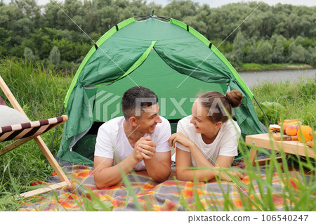 Happy young family resting near the river lying on blanker near the tent talking looking at each other wearing white casual style t-shirts camping adventure trip. Happy young family resting near the river lying on blanker near the tent talking looking at each other wearing white casual style t-shirts camping adventure trip. 106540247