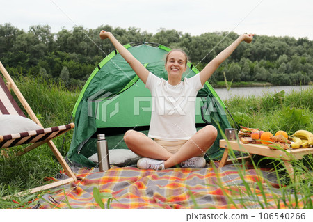 Cheerful woman wearing white t shirt sitting near the tent with crossed legs, raised arms, rejoicing active weekend on nature, looking at camera. 106540266