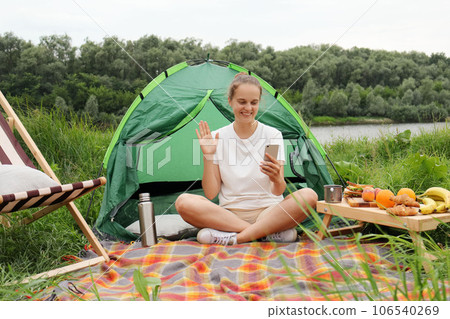 Beautiful woman sitting near tent and river, active lifestyle, traveling, spending time near river, holding smart phone, having video call, waving hello or bye, greeting. 106540269