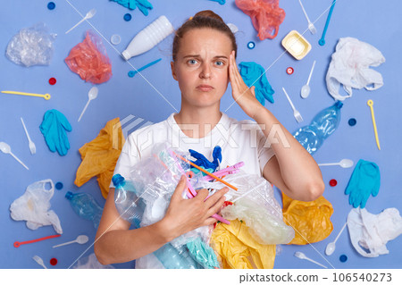Sad unhappy woman wearing white shirt isolated on blue wall with litter around cleaning territory from rubbish looking at camera with displeased expression. Sad unhappy woman wearing white shirt isolated on blue wall with litter around cleaning territory from rubbish looking at camera with displeased expression. 106540273