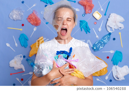 Despair sad unhappy woman wearing white shirt isolated on blue wall with rubbish around holding trash cleaning parks screaming from shock and sorrow environmental pollution. Despair sad unhappy woman wearing white shirt isolated on blue wall with rubbish around holding trash cleaning parks screaming from shock and sorrow environmental pollution. 106540288