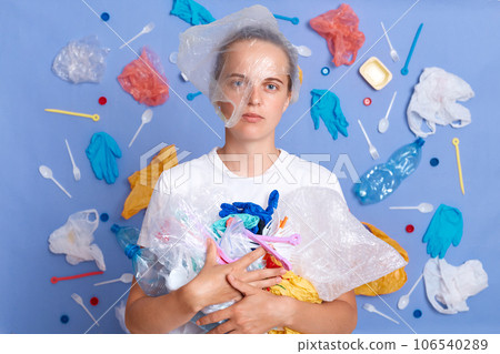 Reuse for eco. Volunteer for cleanup. Protect our planet. Displeased upset woman wearing white shirt isolated on blue wall with garbage around looking at camera with serious face Reuse for eco. Volunteer for cleanup. Protect our planet. Displeased upset woman wearing white shirt isolated on blue wall with garbage around looking at camera with serious face 106540289