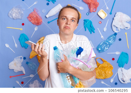 Uncertain woman wearing white shirt isolated on blue wall with garbage around standing with litter and smart phone in hands looking at camera shrugging shoulders. 106540295