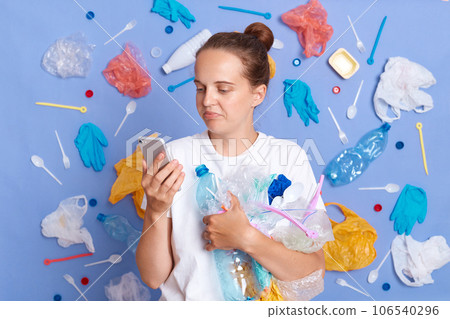 Brown haired woman wearing white shirt isolated on blue wall with rubbish around using mobile phone, reading messages i n social networks, having break while cleaning up street from trash. 106540296