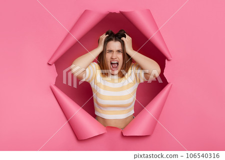 Irritated frustrated brunette woman wearing striped top looking through ripped pink paper hole being in panic pulling her hair screaming with anger. Irritated frustrated brunette woman wearing striped top looking through ripped pink paper hole being in panic pulling her hair screaming with anger. 106540316