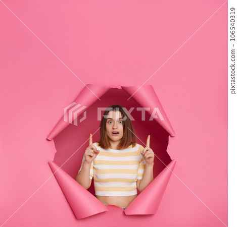 Shocked scared brunette woman wearing striped top looking through ripped pink paper hole pointing upwards looking with big eyes pointing at empty space for advertisement. 106540319
