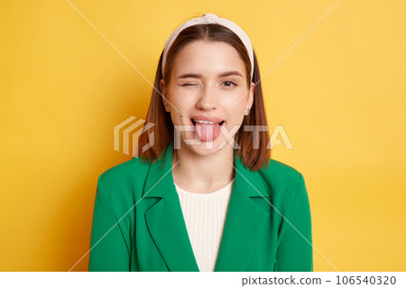 Childish behavior. Playful positive woman wearing green jacket winking to camera showing tongue out posing isolated over yellow background. 106540320