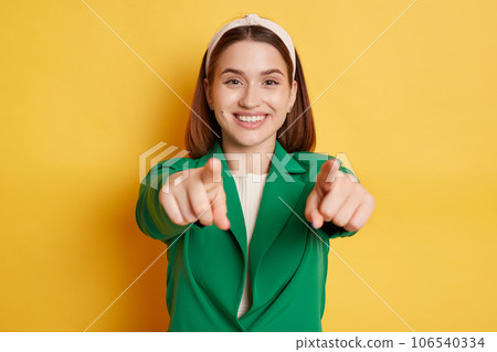 Joyful smiling woman wearing green jacket posing isolated over yellow background pointing to camera choosing you positive reaction. Joyful smiling woman wearing green jacket posing isolated over yellow background pointing to camera choosing you positive reaction. 106540334