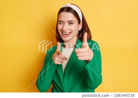 Flirting positive woman wearing green jacket posing isolated over yellow background showing finger guns to camera winking expressing happiness. 106540336