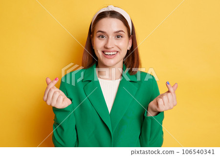 Excited amazed woman wearing green jacket posing isolated over yellow background standing showing korean love gesture expressing love. Excited amazed woman wearing green jacket posing isolated over yellow background standing showing korean love gesture expressing love. 106540341