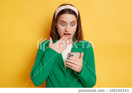 Shocked woman wearing green jacket posing isolated over yellow background using mobile phone reading news in social networks. Shocked woman wearing green jacket posing isolated over yellow background using mobile phone reading news in social networks. 106540343
