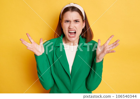 Angry irritated woman wearing green jacket posing isolated over yellow background arguing screaming with hate raised her arms yelling with anger. 106540344