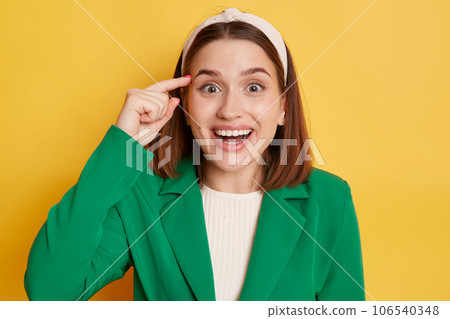 Surprised amazed woman wearing green jacket standing isolated over yellow background looking at camera with open mouth and big eyes showing stupid gesture. Surprised amazed woman wearing green jacket standing isolated over yellow background looking at camera with open mouth and big eyes showing stupid gesture. 106540348