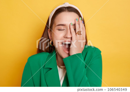 Positive laughing woman wearing green jacket standing isolated over yellow background showing facepalm covering half of face with hand closed eyes. Positive laughing woman wearing green jacket standing isolated over yellow background showing facepalm covering half of face with hand closed eyes. 106540349