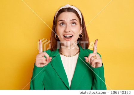 Surprised amazed woman wearing green jacket posing isolated over yellow background pointing up at copy space for promotion advertisement area. Surprised amazed woman wearing green jacket posing isolated over yellow background pointing up at copy space for promotion advertisement area. 106540353