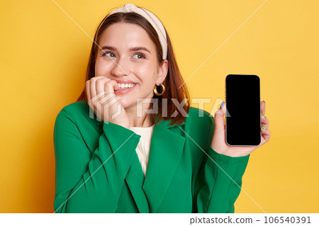 Dreaming smiling satisfied woman wearing green jacket posing isolated over yellow background showing cell phone advertisement empty space for promotion. 106540391