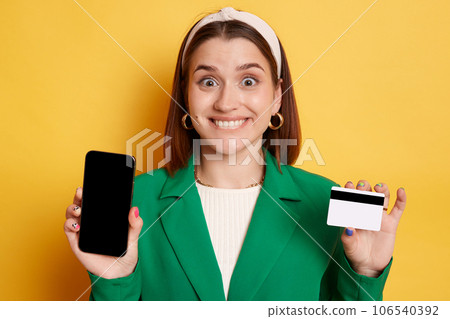 Phone banking. Online payments. Delighted happy woman wearing green jacket posing isolated over yellow background showing mobile phone with empty display and credit card. 106540392