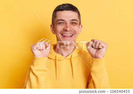 Joyful happy smiling man wearing casual hoodie clenched fists celebrating success rejoicing his achievements posing isolated over yellow background. Joyful happy smiling man wearing casual hoodie clenched fists celebrating success rejoicing his achievements posing isolated over yellow background. 106540455
