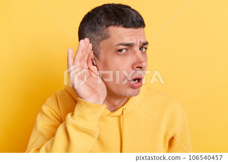 Confused man wearing casual hoodie standing with hands near ear trying to listen silent talking posing isolated over yellow background. Confused man wearing casual hoodie standing with hands near ear trying to listen silent talking posing isolated over yellow background. 106540457