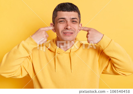 Despair sad man wearing casual hoodie covering mouth with fingers hearing loud noise frowning face posing isolated over yellow background. Despair sad man wearing casual hoodie covering mouth with fingers hearing loud noise frowning face posing isolated over yellow background. 106540458